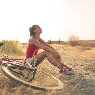 Full body of female in shorts and top sitting on roadside in rural field with bicycle near and enjoying fresh air with eyes closed