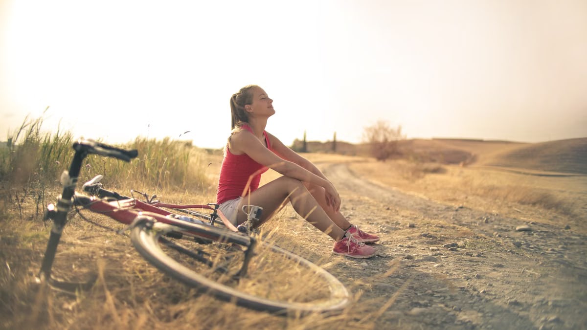 Full body of female in shorts and top sitting on roadside in rural field with bicycle near and enjoying fresh air with eyes closed